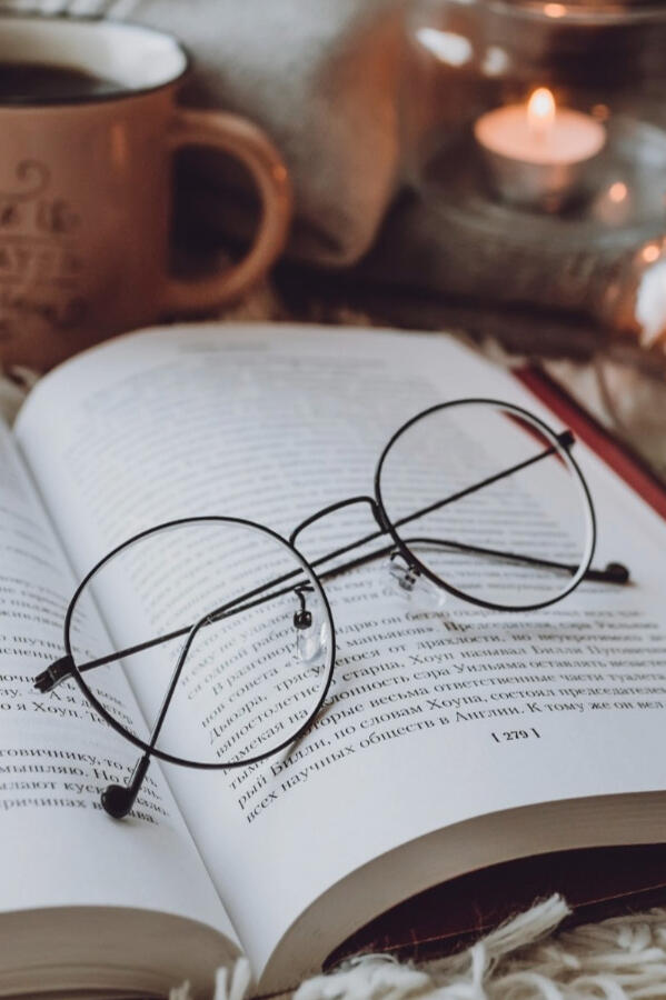 Cosy reading scene with an open book, round glasses resting on the pages, a warm drink in a ceramic mug, lit tealight candles in glass holders, and a folded sweater in the background.